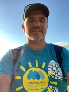 Man in cap with Miscarriage Association Miles That Matter t-shirt on, with blue sky behind him.