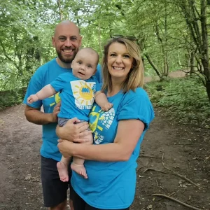Katrina, her partner, and baby Harry smiling together in matching blue Miscarriage Association t-shirts during their Miles That Matter walk in a wooded area.