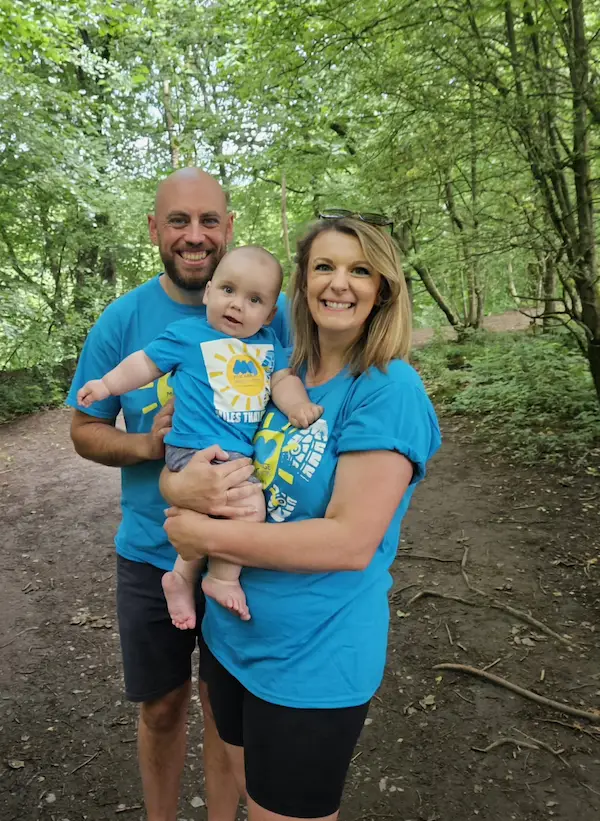 Katrina, her partner, and baby Harry smiling together in matching blue Miscarriage Association t-shirts during their Miles That Matter walk in a wooded area.