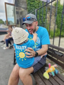 Adam holding and smiling at baby Harry, both wearing matching blue ‘Miles That Matter’ t-shirts during their 2025 fundraising challenge